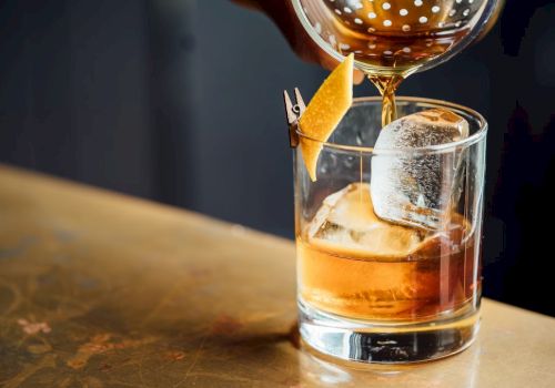 A glass of whiskey with a large ice cube and orange peel garnishes, being poured from a cocktail shaker, on a wooden bar.