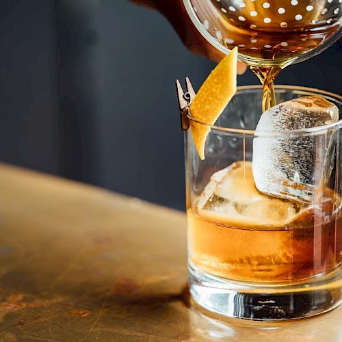 A glass of whiskey with a large ice cube and orange peel garnishes, being poured from a cocktail shaker, on a wooden bar.