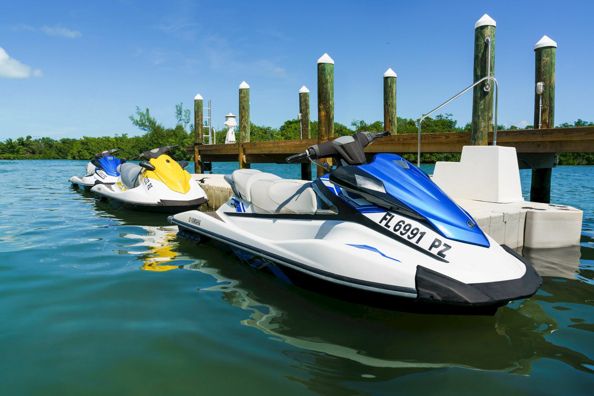 Several jet skis docked at a wooden pier on calm blue water; clear sky, green vegetation in the distance, and numbered registration on the closest jet ski.