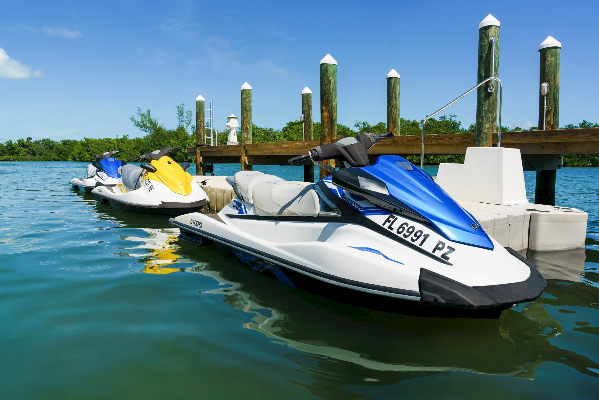 Several jet skis docked at a wooden pier on calm blue water; clear sky, green vegetation in the distance, and numbered registration on the closest jet ski.