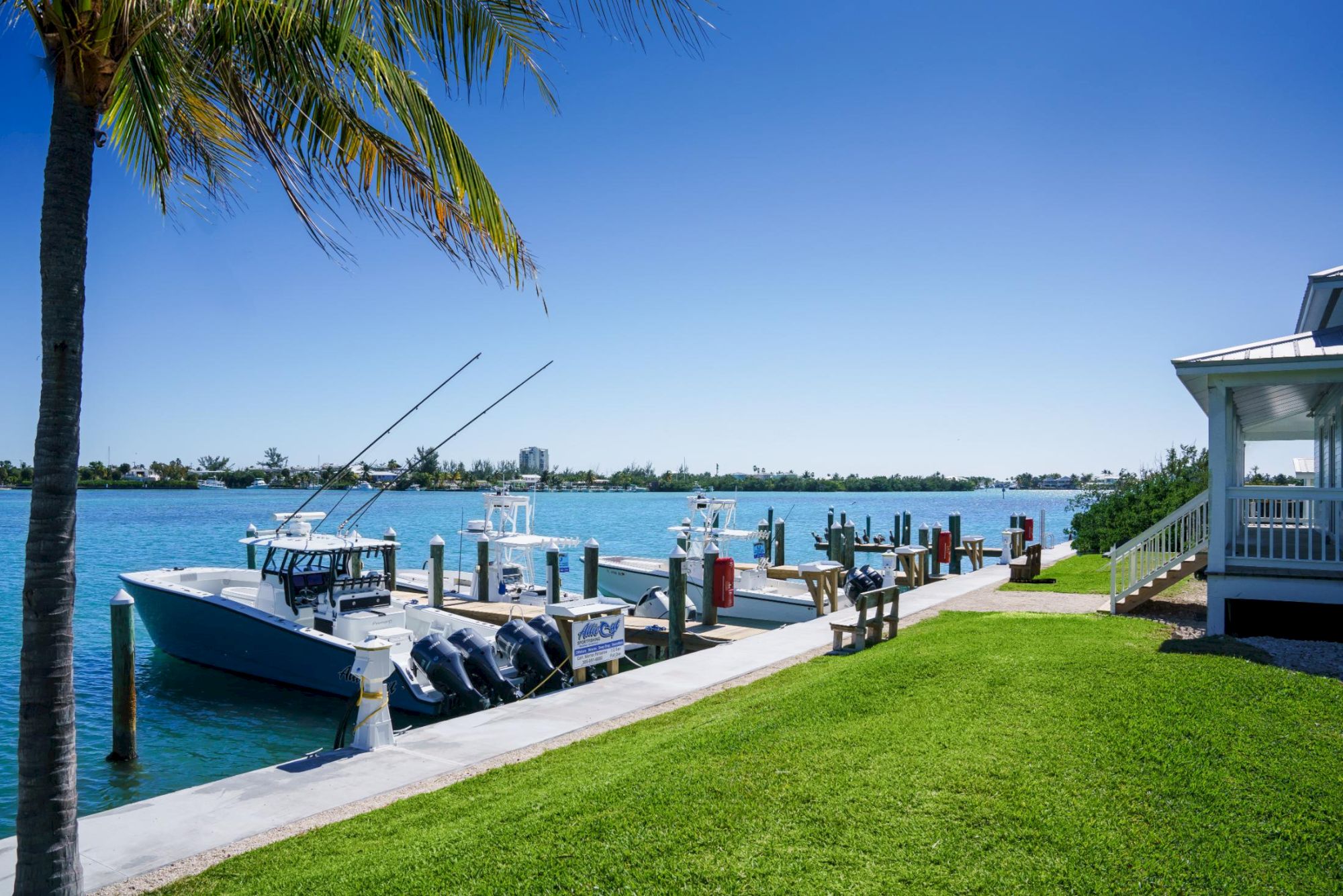 A sunny marina with a docked boat, fishing rods, palm trees, bright blue water, a grassy shoreline, and a white dockside building, perfect for a relaxing day.