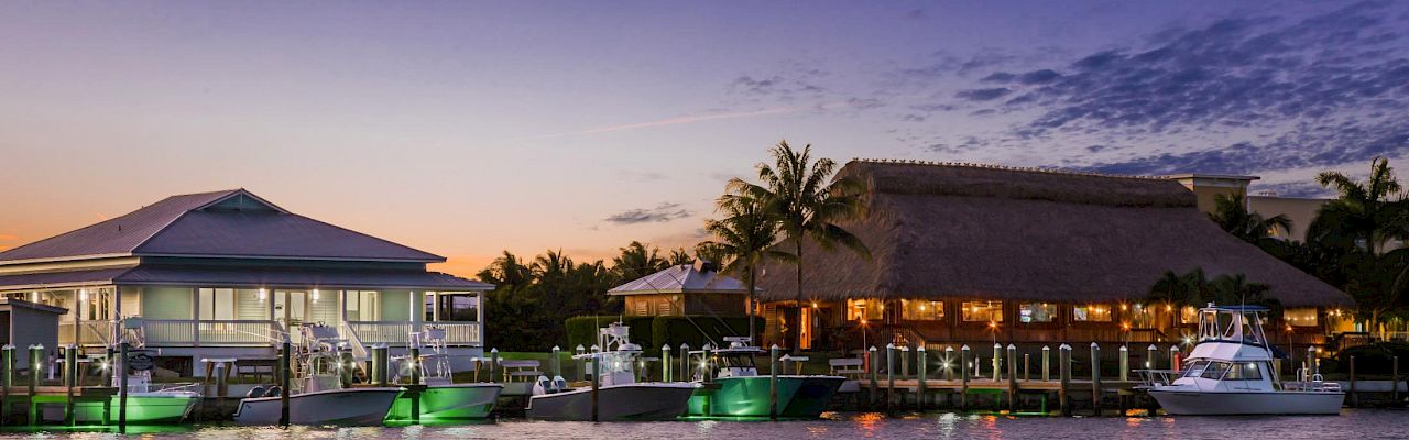 A tranquil waterfront at dusk with palm trees, thatched-roof huts, and boats docked along a lit pier, reflecting calm waters.