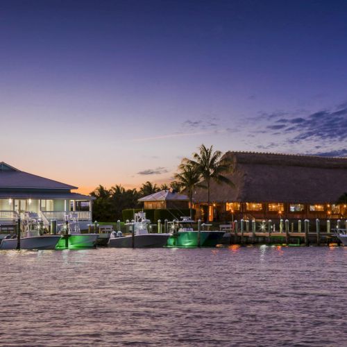 A tranquil waterfront at dusk with palm trees, thatched-roof huts, and boats docked along a lit pier, reflecting calm waters.