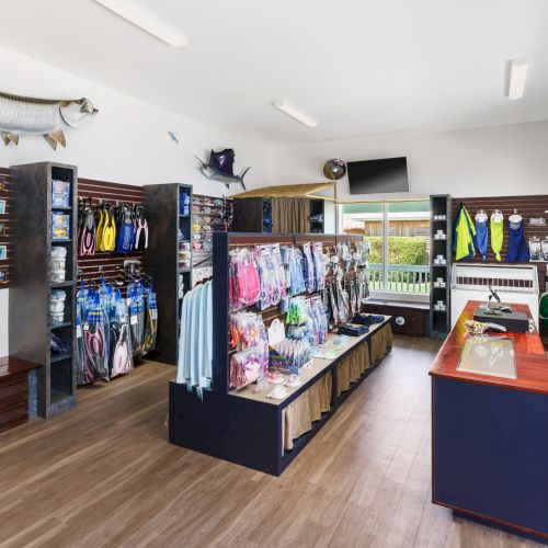A fishing/outdoor gear shop with clothing racks, waders, tackle, kayaks, and a display counter; mounted fish decor on the wall.