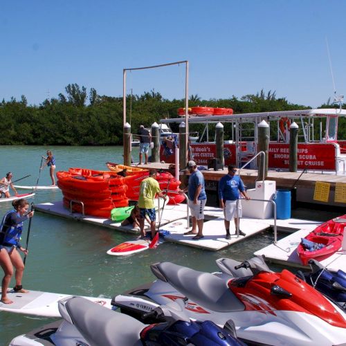 Bystanders watch as a crowded boat rescue scene on a turquoise sea, with people in orange life vests near a small vessel and onlookers ashore.