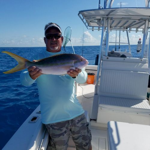 A man on a boat, holding a fish he just caught, with blue ocean and clear sky in the background, smiling proudly.