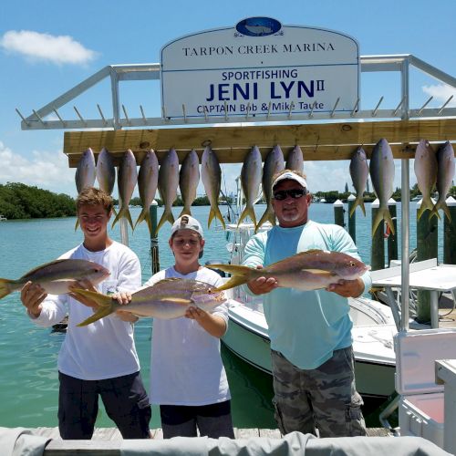 A group of smiling people pose with fishing rods on a dock, in front of a building marked &ldquo;Lodges&rdquo; and a blue sky.
