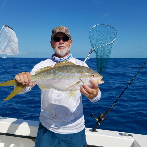 A man wearing sunglasses and a blue shirt on a boat, holding a large fish he just caught, with a clear blue sky and ocean in the background.