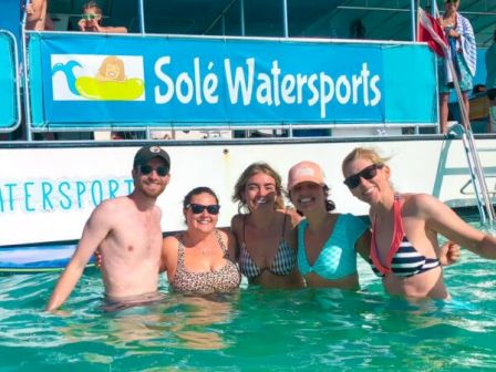 Six friends posing in a pool at a Sol&eacute; Watersports event, smiling and wearing swimsuits, with a banner in the background.