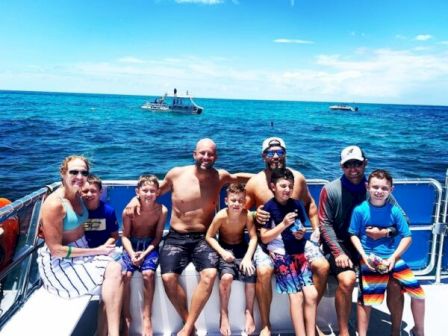 A group of eight people (mostly kids) in swimsuits sit on a boat railing, smiling with the ocean and other boats behind them.