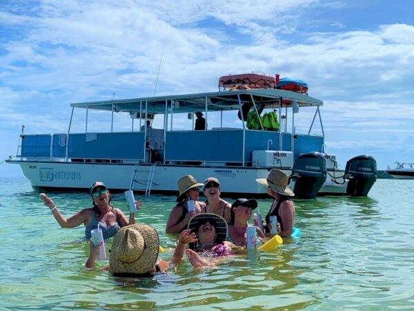 A group of people in shallow turquoise water posing for a photo near a blue boat with a covered upper deck at a sunny beach.