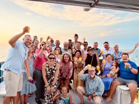 A large group of friends on a boat, posing for a selfie, many smiling and raising drinks as the sunset skies glow above.