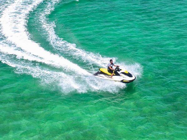 A person rides a yellow and black jet ski on clear, turquoise water, creating white frothy waves as they speed along, sunny day.