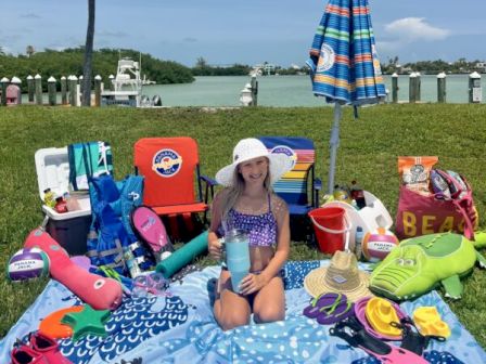 A young girl sits on a blue beach blanket with water toys, towels, and bags spread around, near a lake with a striped umbrella and chairs.