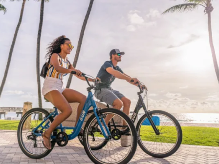 Two people ride blue bicycles along a sunny beach path with palm trees in the background, wearing casual summer clothes and backpacks, enjoying a ride.