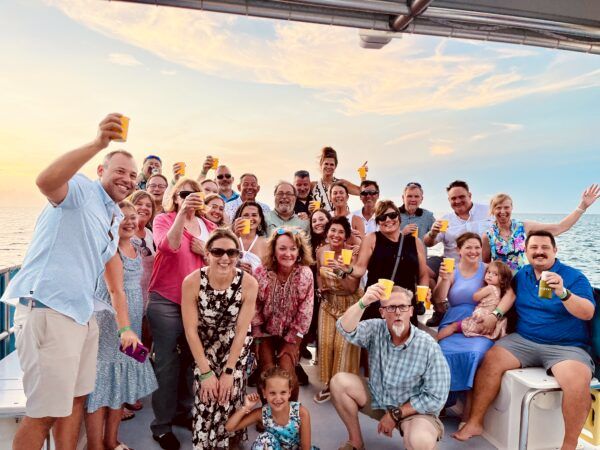 A large group of friends on a boat party, posing for a selfie while cheering with drinks and smiling under a sunny sky.