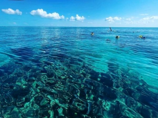 Clear turquoise sea with transparent water, sandy bottom visible, a few distant divers, and a bright blue sky with white clouds ending the sentence.