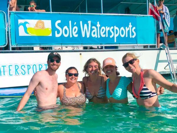 Six friends posing in a pool at a Sol&eacute; Watersports event, smiling and wearing swimsuits, with a banner in the background.