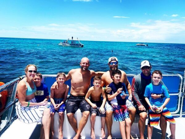 A group of eight people (mostly kids) in swimsuits sit on a boat railing, smiling with the ocean and other boats behind them.