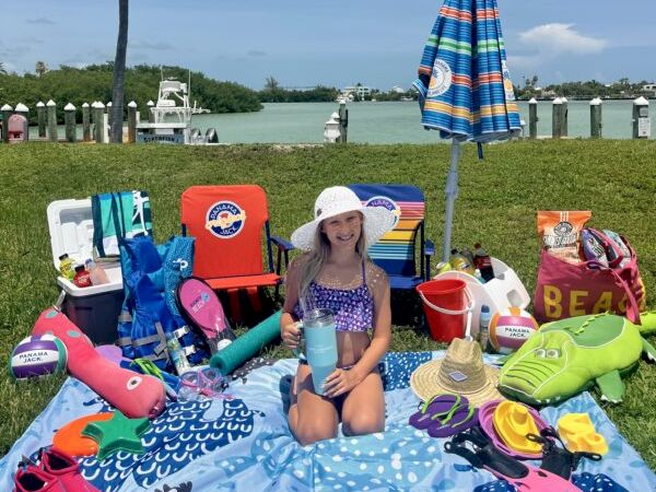 A young girl sits on a blue beach blanket with water toys, towels, and bags spread around, near a lake with a striped umbrella and chairs.