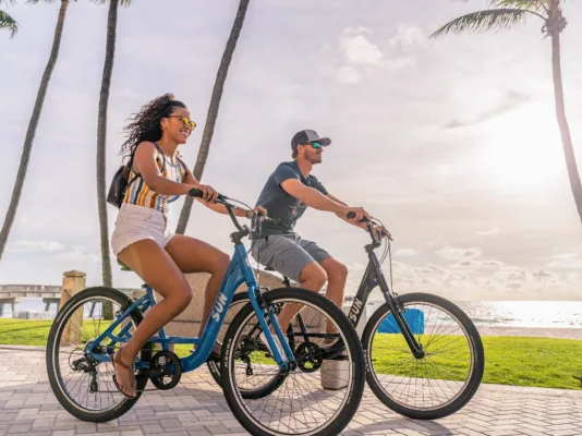 Two people ride blue bicycles along a sunny beach path with palm trees in the background, wearing casual summer clothes and backpacks, enjoying a ride.