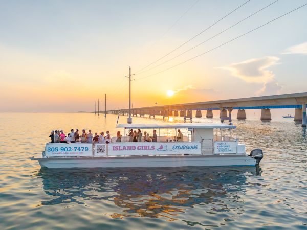 A sunset cruise boat with passengers floats on calm water near long pier pylons, with the &ldquo;Island Girls Excursions&rdquo; sign visible.