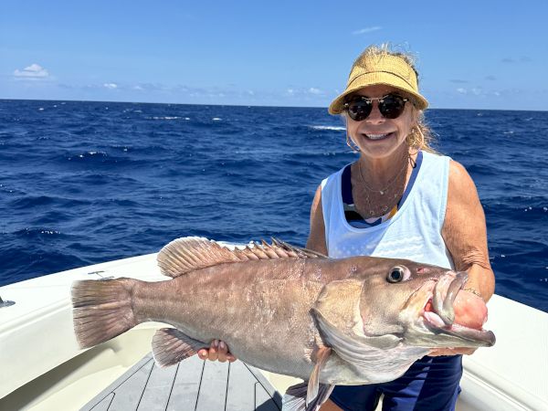 On a sunny boat, a woman in a hat and sunglasses proudly holds up a big caught fish with the ocean behind her.