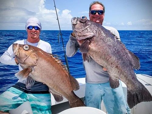 Two anglers on a boat pose with big ocean fish they caught in open water, smiling under a bright blue sky.