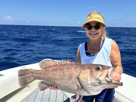 On a sunny boat, a smiling woman holds up a big fish while fishing in the open ocean.